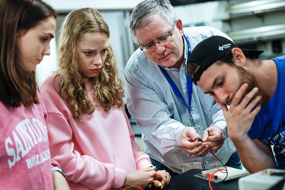 An instructor working with a group of three students. The students look on as the instructor holds a small electric device in his hands