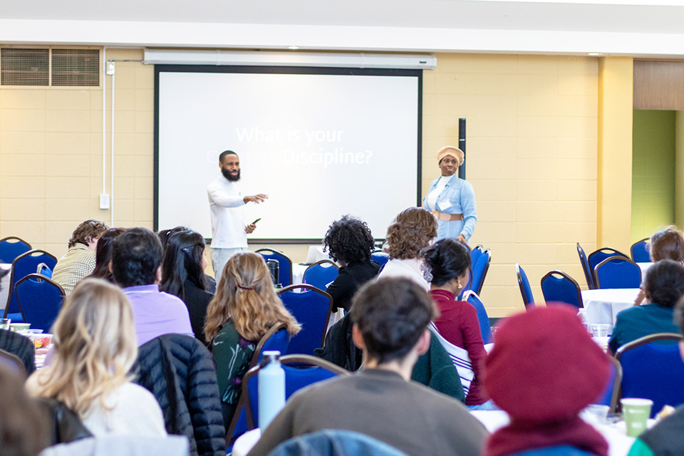 Two presenters speak while the audience, seen from behind, looks on.