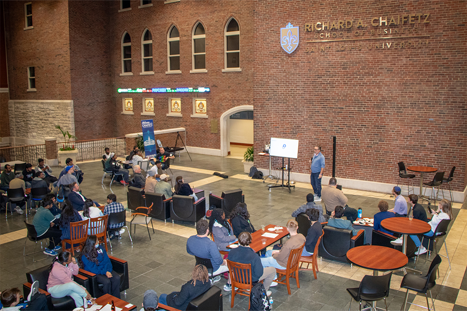 People sit around tables in an atrium while a speaker holds a microphone at one end of the room.