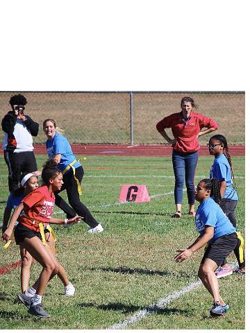 Students play on a soccer field while adults observe in the background.