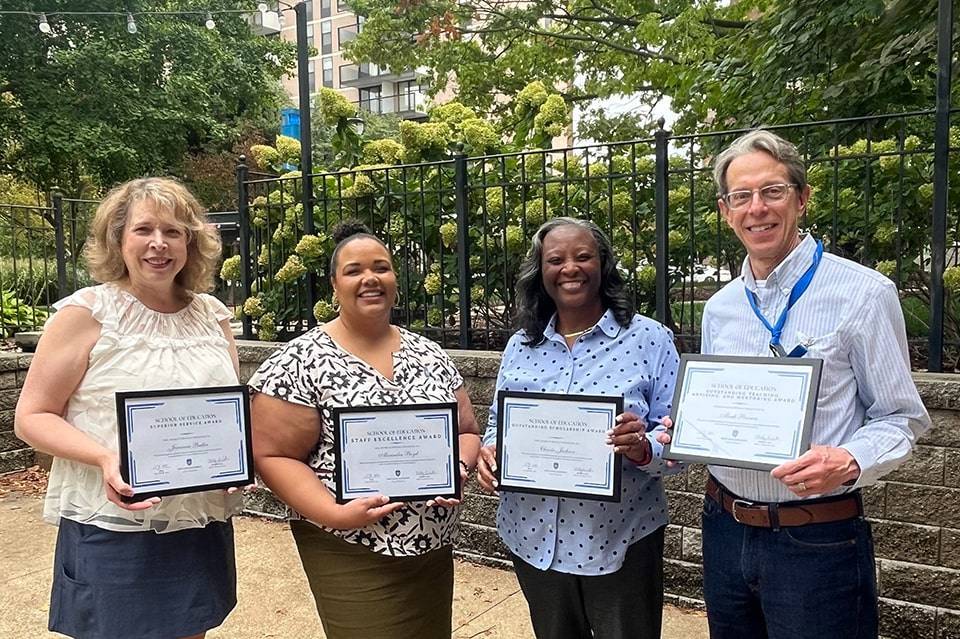 Faculty staff awards Four people hold awards plaques while standing under a tree.