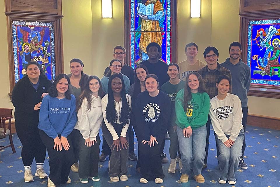 A group of 15 SLU students and their staff supervisor pose for a photo in a room at DuBourg Hall with stained glass windows behind them.