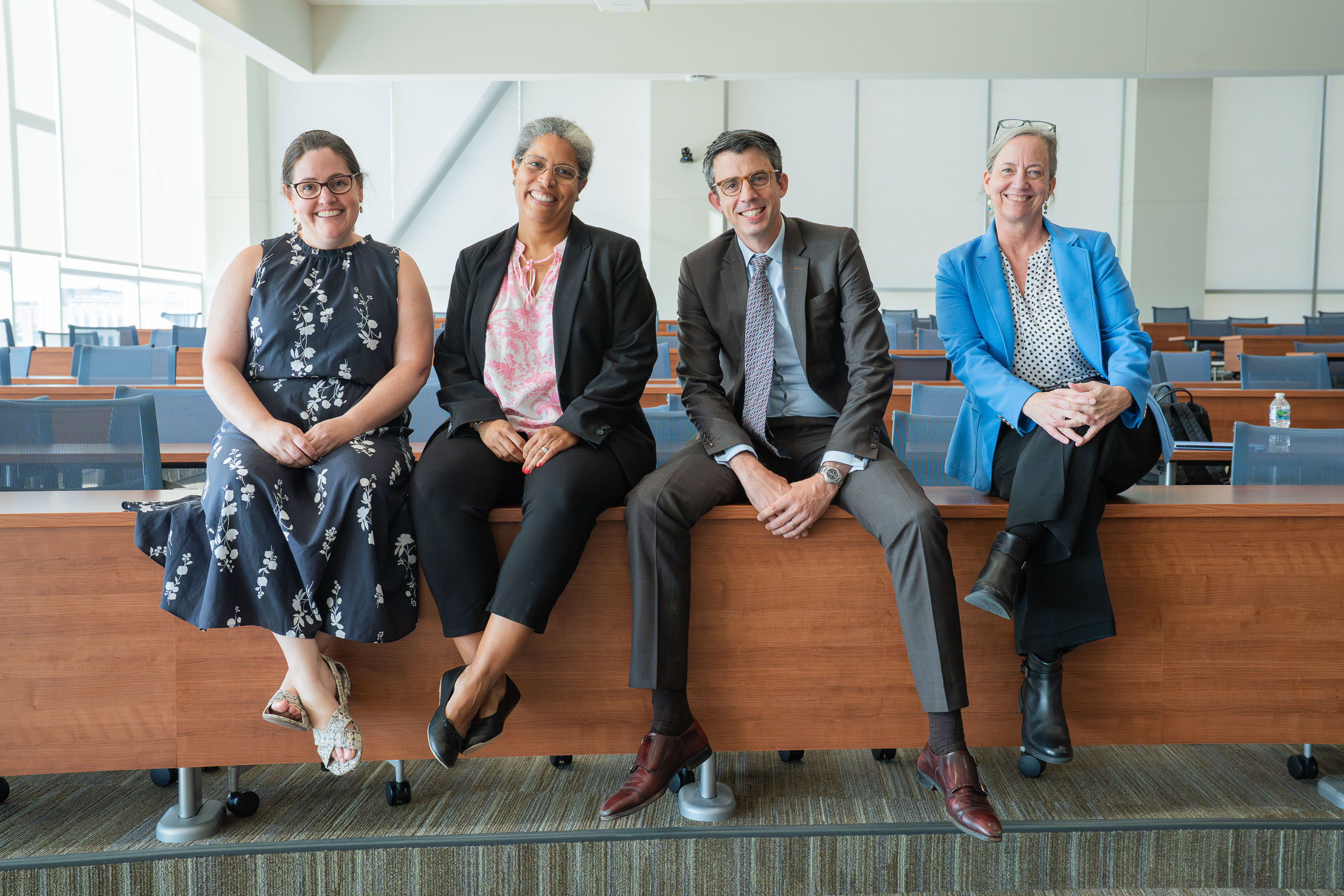 Four faculty members sit on a desk in a lecture hall.