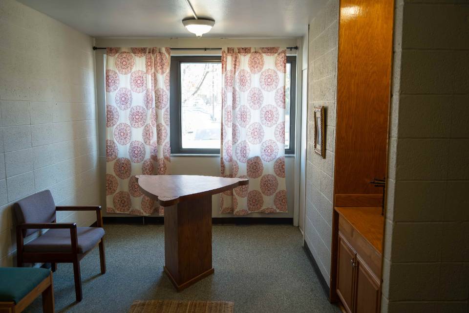 A room with a triangular table in the center, a few chairs and white curtains with a floral pattern.