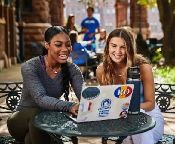 Two smiling students sitting outdoors at a small cafe table studying with laptops in front