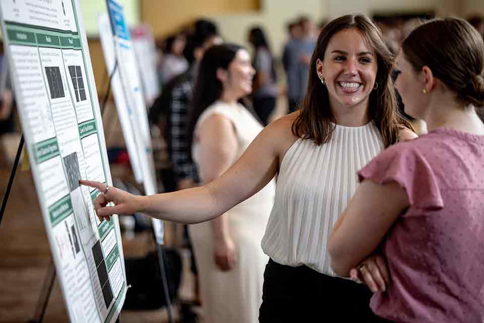 A senior smiles as she shows off her research poster to an attendee of the 2024 Senior Legacy Symposium