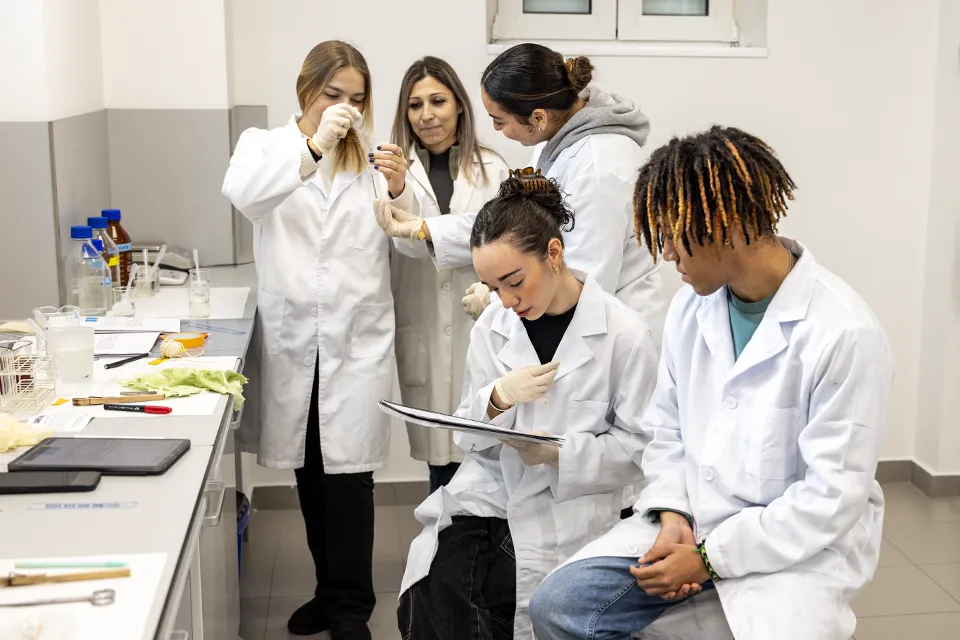 A group of students with their professor, wearing white coats, in a biology lab.