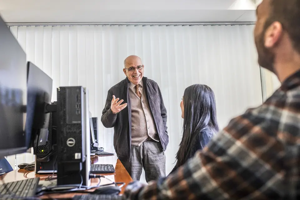 Two students sitting in front of their computers, attending to a teacher in the background of the image.