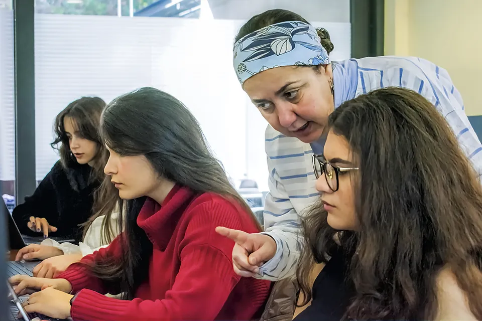 A teacher points at a laptop screen while a student pays attention. Two students look at their laptops in the background.