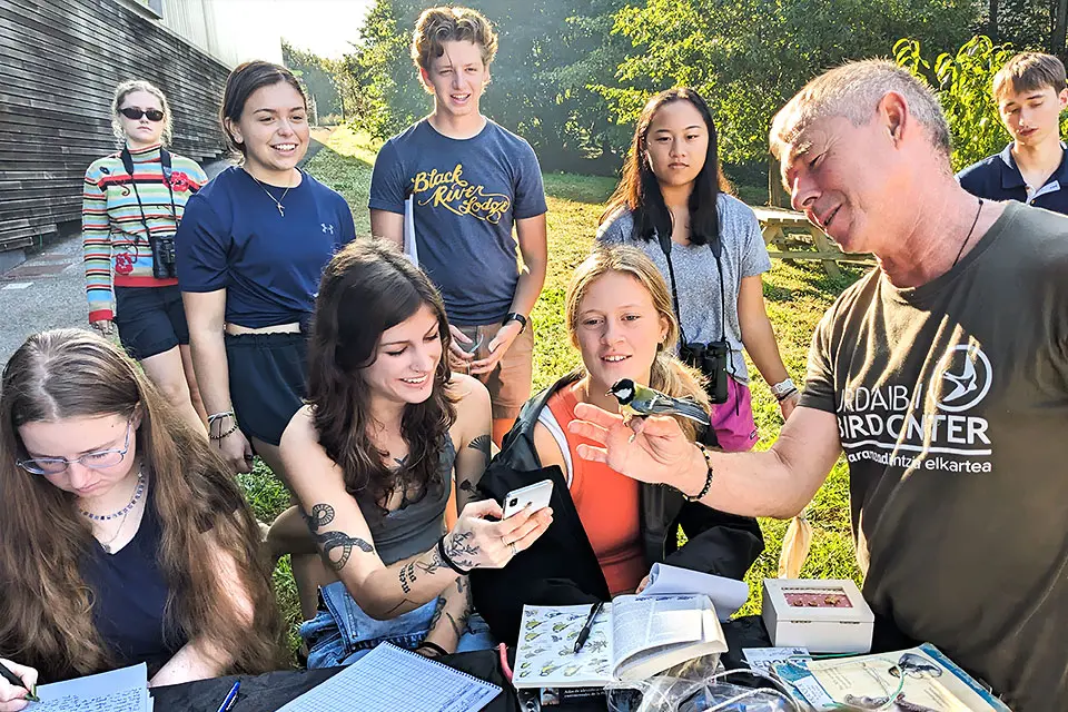 A group of students, in an outdoor class, look intently at a teacher holding a bird on his hand.