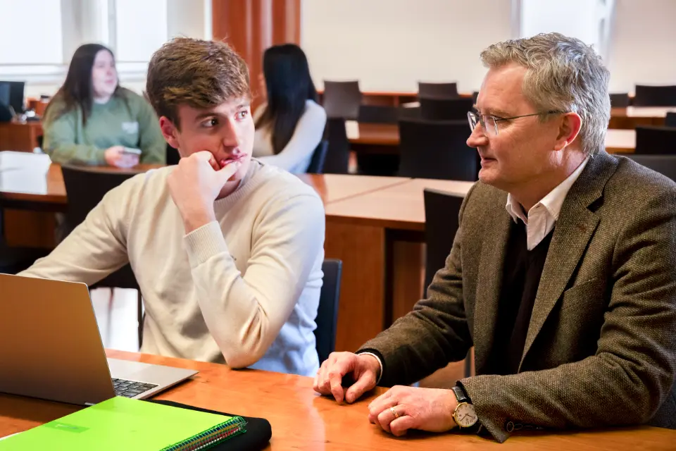 A student sitting at a table with his laptop and a professor in a suit next to him.