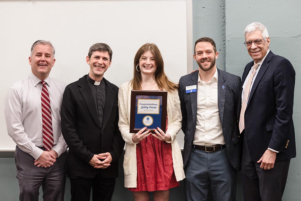 Gabby Chiodo holds her plaque while standing at the front of a classroom, between Provost Mike Lewis, Father Matthew Baugh, Dr. Robert Pampel and SLU President Dr. Fred Pestello.