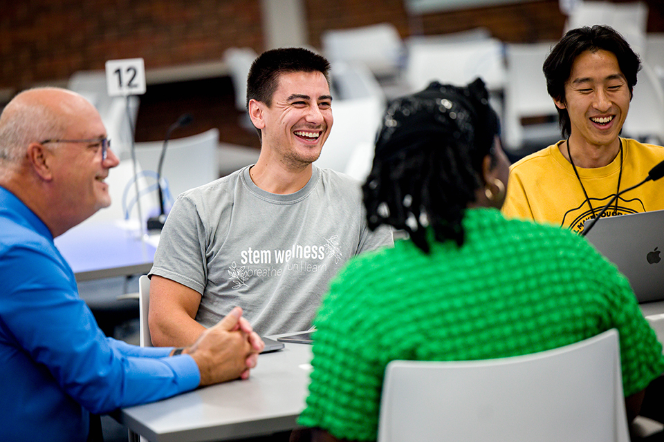 A group of four people sit at a table in an interprofessional education course. They are smiling and laughing.