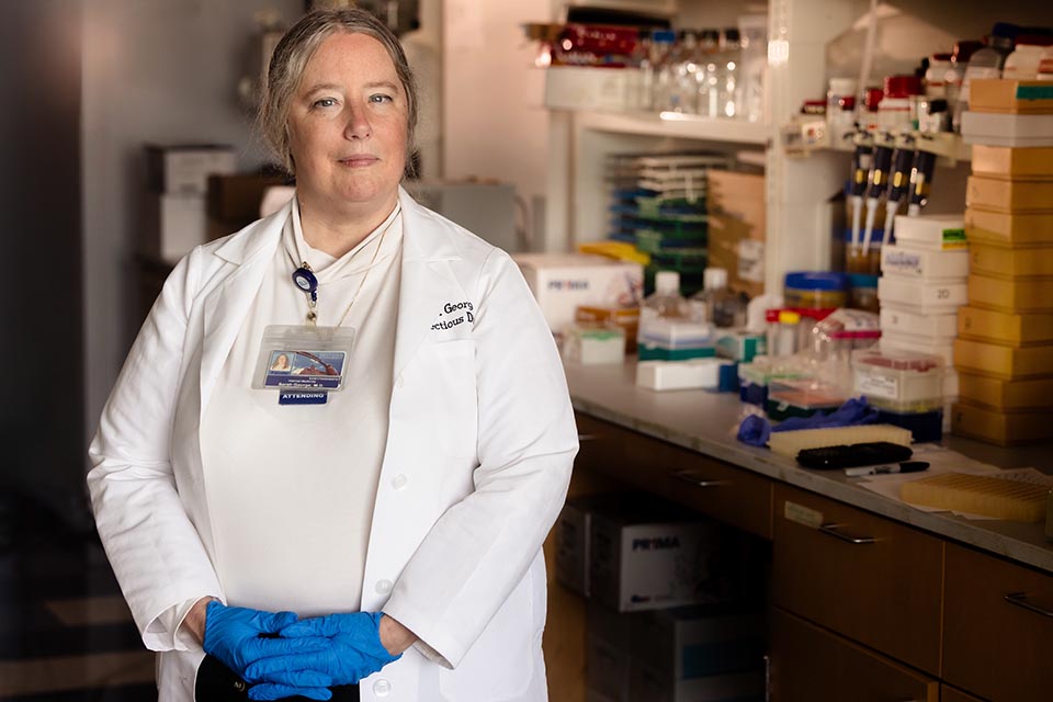 Sarah George, M.D., poses for a photo inside a dimly lit lab. She is wearing a white shirt, a name tag, a white lab coat, and blue latex gloves. Her hands are interlocked at the waist. 