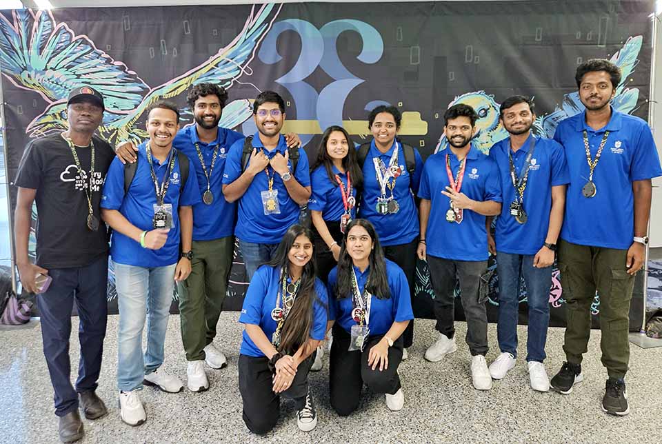A group wearing blue shirts and medals around their necks pose for a photo. A man on the far left wears a black shirt and hat and is also wearing a medal around his neck.