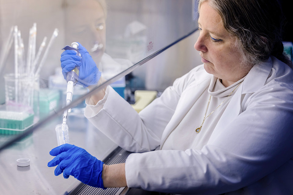 Dr. Sarah George holds a syringe over a tube in a lab. She is wearing a white lab coat and blue gloves.