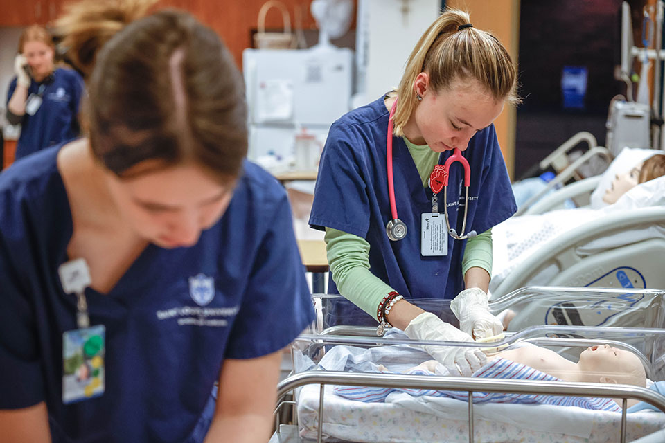 Up close image of two nursing students in the simulation lab. One of the students is changing the diaper of a baby mannequin. Both students are wearing Saint Louis University blue nursing scrubs. A third student can be seen in the background on the telephone.