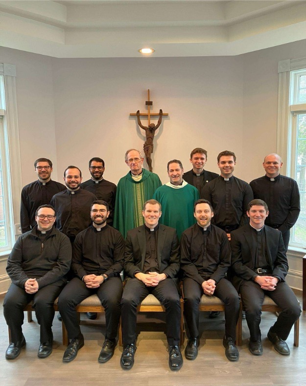 A group of Jesuits in formation gathered in a religious ceremony, receiving their ministry assignments. They stand in a solemn setting, dressed in traditional clerical attire, symbolizing their commitment to service and faith.