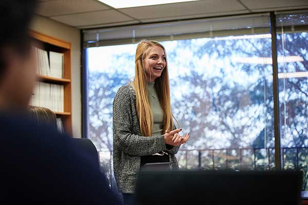 A student in a green and grey sweater stands at a conference table and presents to their peers in a big meeting room with a large window.