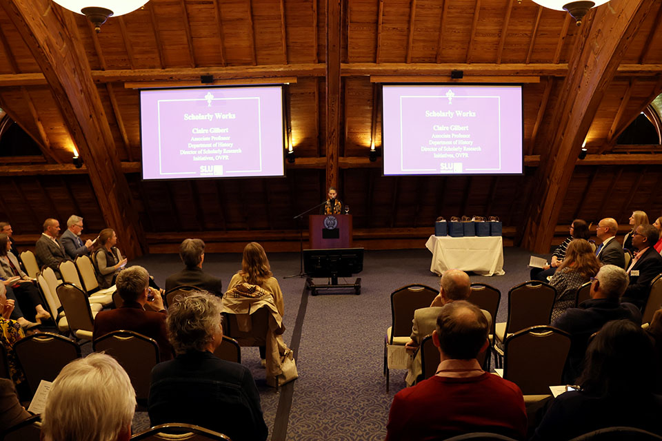 A woman stands at a lectern in front of an audience, with a video screen on each side