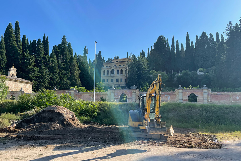 A piece of earth-moving equipment sits on a pile of dirt, with an ornate wall and home on a hillside in the background.