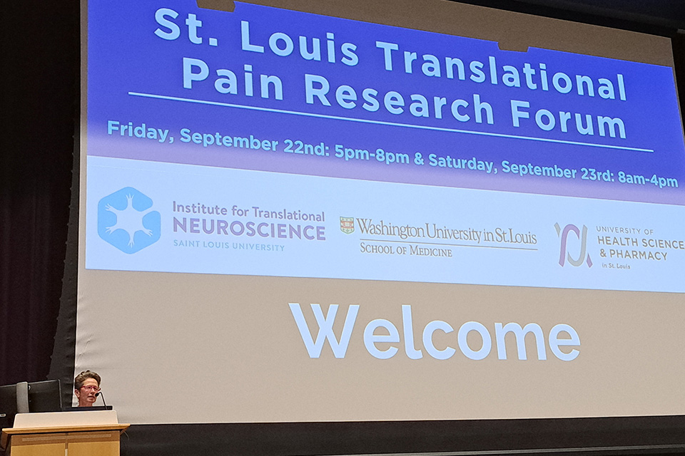 A speaker stands at a podium below a large screen that reads St. Louis Translational Pain Research Forum, Friday, September 22nd: 5 p.m.-8 p.m., and Saturday, September 23rd, 8 a.m. to 4 p.m. The screen also contains logos for the Institute for Translational Neuroscience, the Washington University School of Medicine and the University of Health Sciences and Pharmacy.