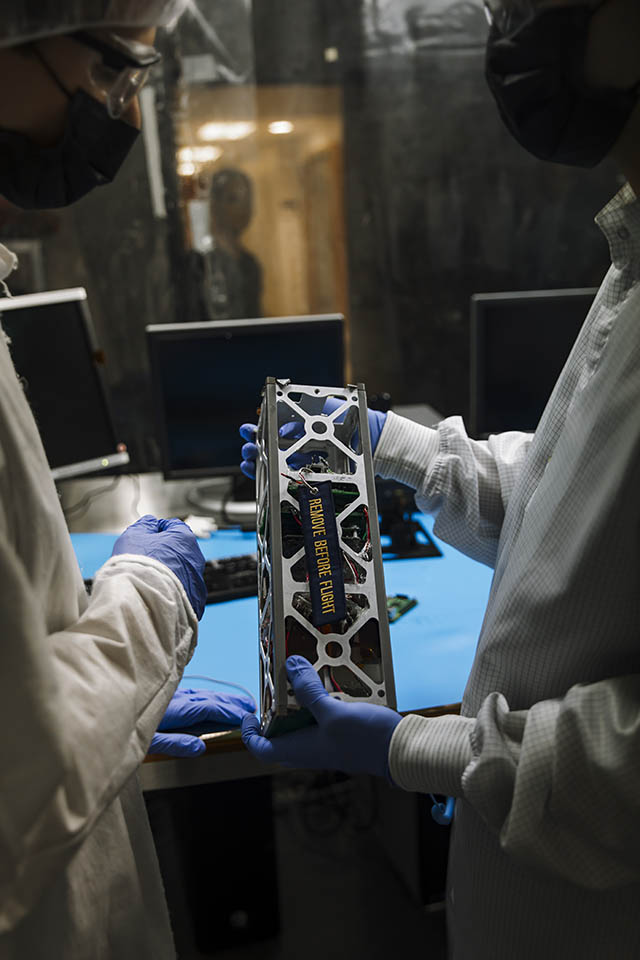 The hands of a student are seen holding a small satellite that has a tag with the words "remove before flight" on it.