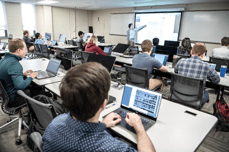 Students sit at long rows of desks in a classroom with laptops in front of them while an instructor points to a presentation screen.