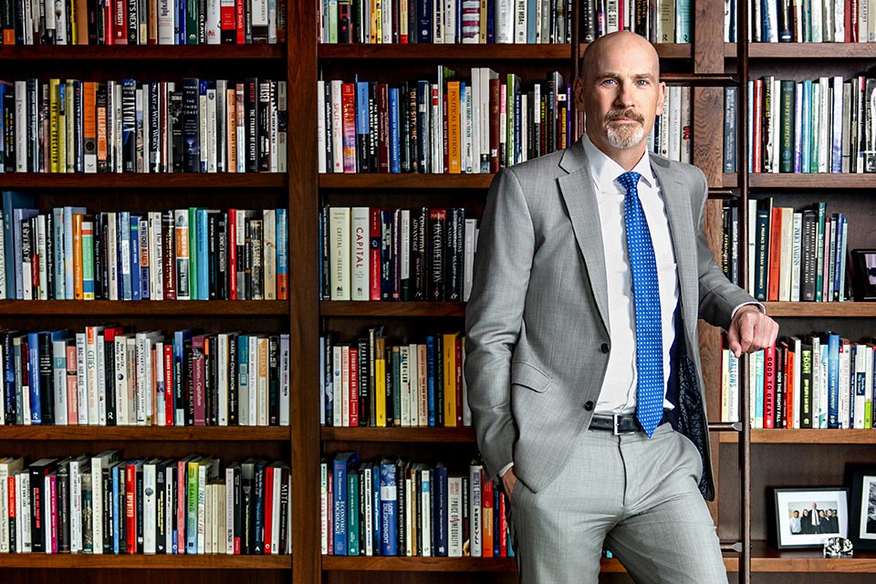 SLU President Dr. Edward Feser A bald white man in a grey suit stands against a wall of books
